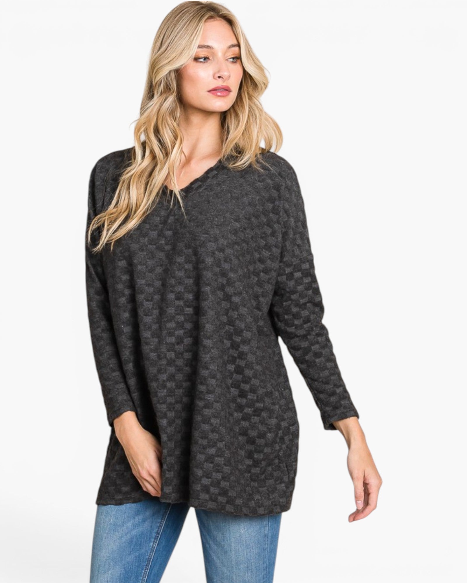 A woman modeling a dark gray long-sleeve knit tunic top with a subtle checkerboard pattern, paired with blue jeans, against a plain white studio background.