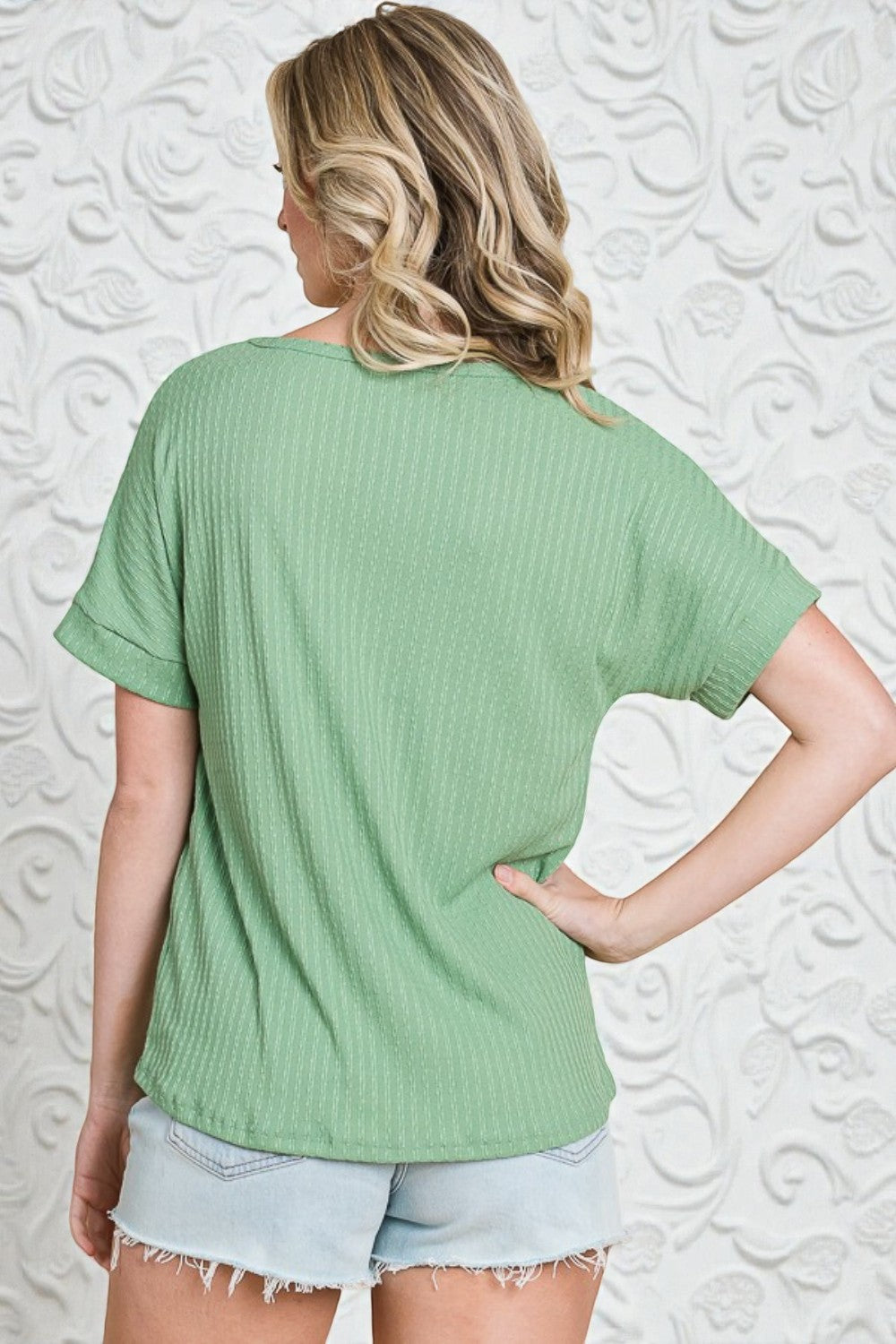 Woman wearing a green ribbed t-shirt and denim shorts against a textured white wall.