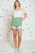 Woman wearing a green and white top with denim shorts against a colorful geometric-patterned wall.