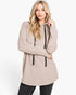 A woman modeling a light beige hooded knit tunic sweater with black drawstrings, paired with black jeans, against a plain white studio background.
