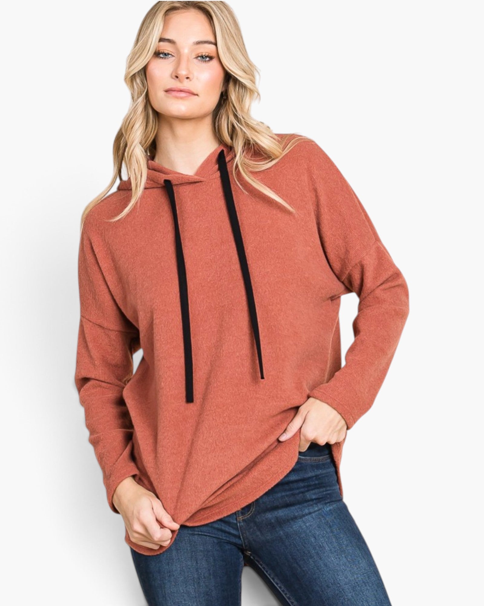 A woman wearing a rust-colored pullover hoodie with black drawstrings and blue jeans, standing against a plain white studio background.