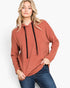 A woman wearing a rust-colored pullover hoodie with black drawstrings and blue jeans, standing against a plain white studio background.