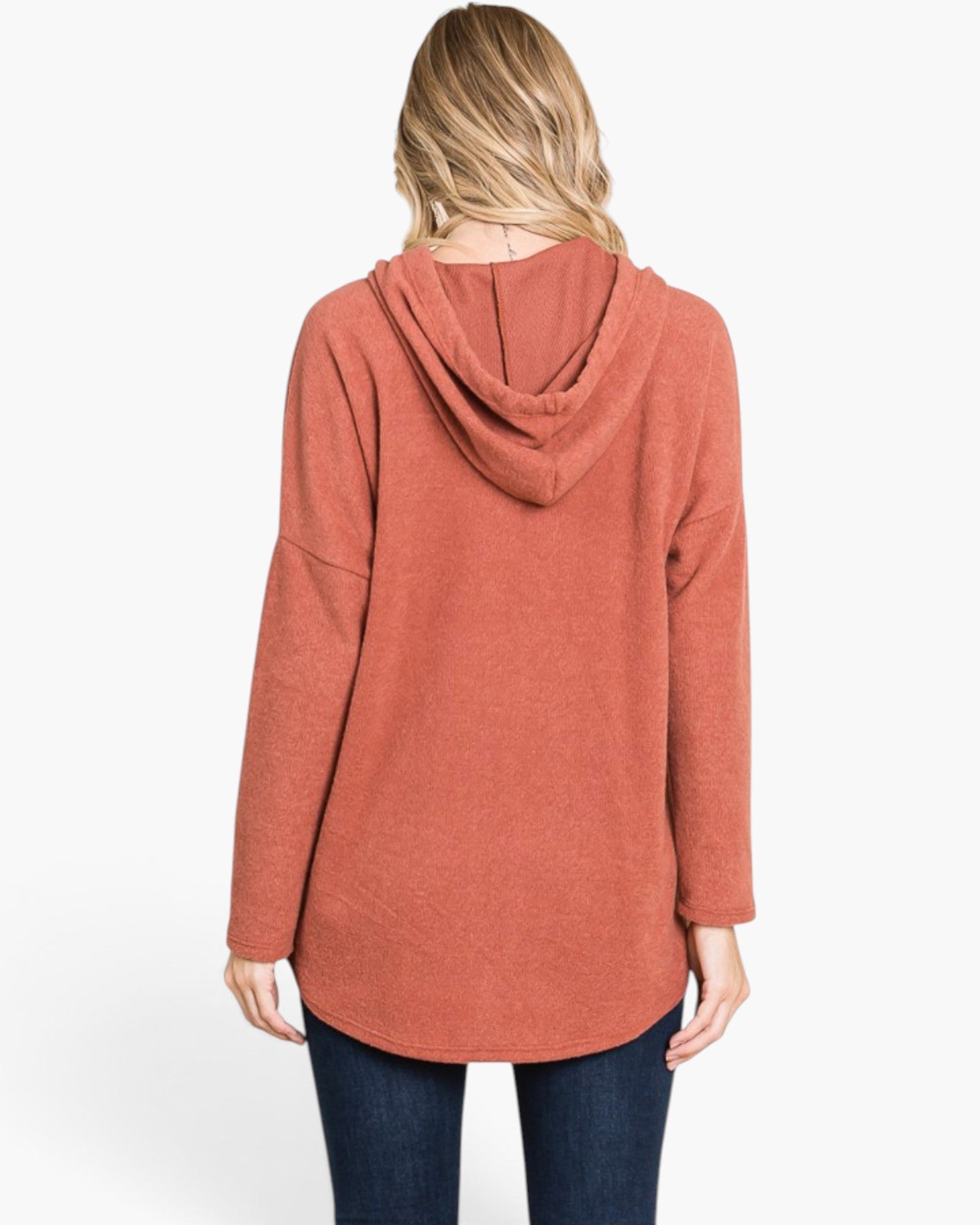 A woman viewed from the back wearing a loose, long-sleeve hooded top in a muted terracotta or burnt orange color, paired with dark blue jeans, against a plain white studio background.
