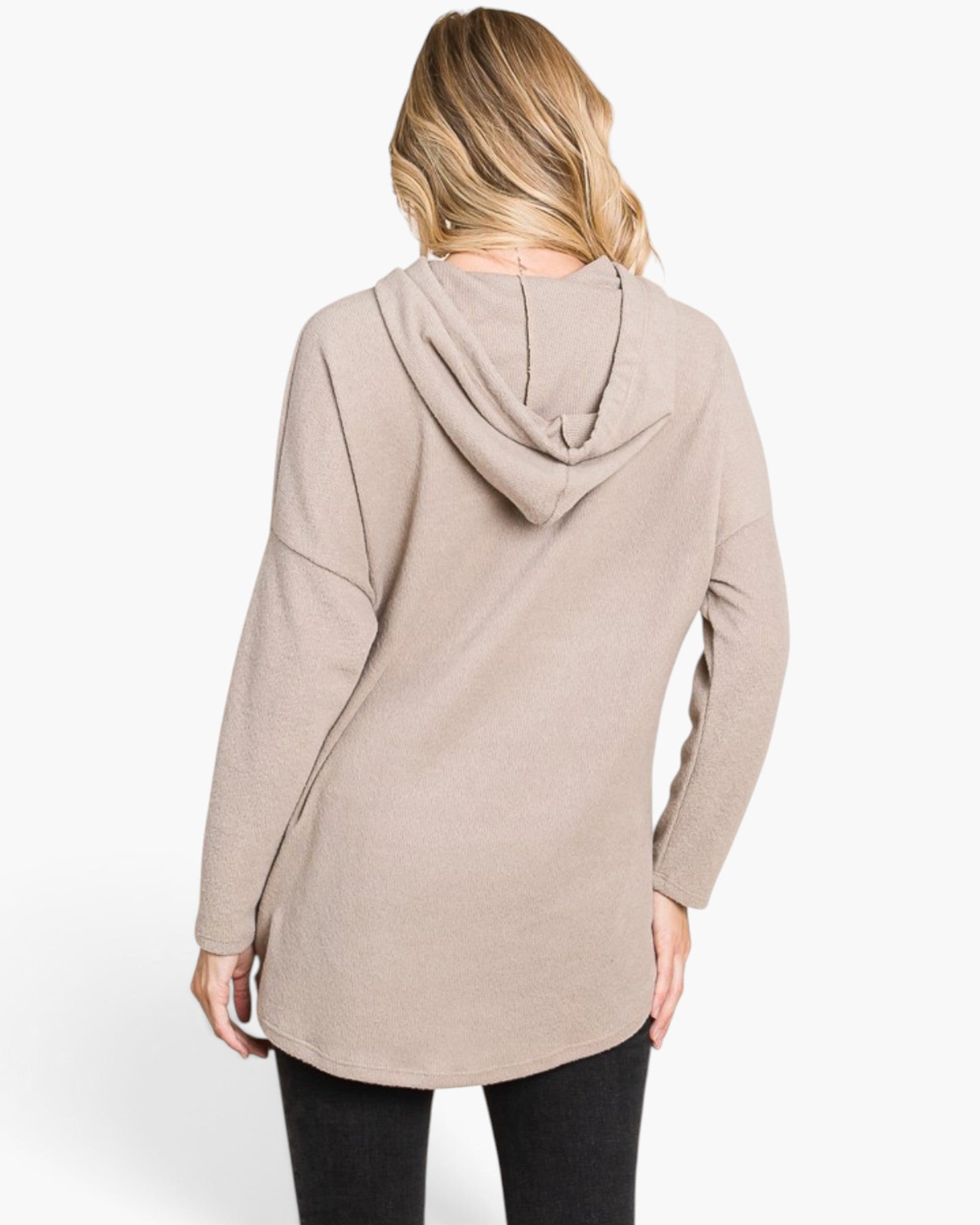 A woman shown from the back wearing a light taupe-beige hooded long-sleeve tunic top and black pants, photographed against a clean white studio background.