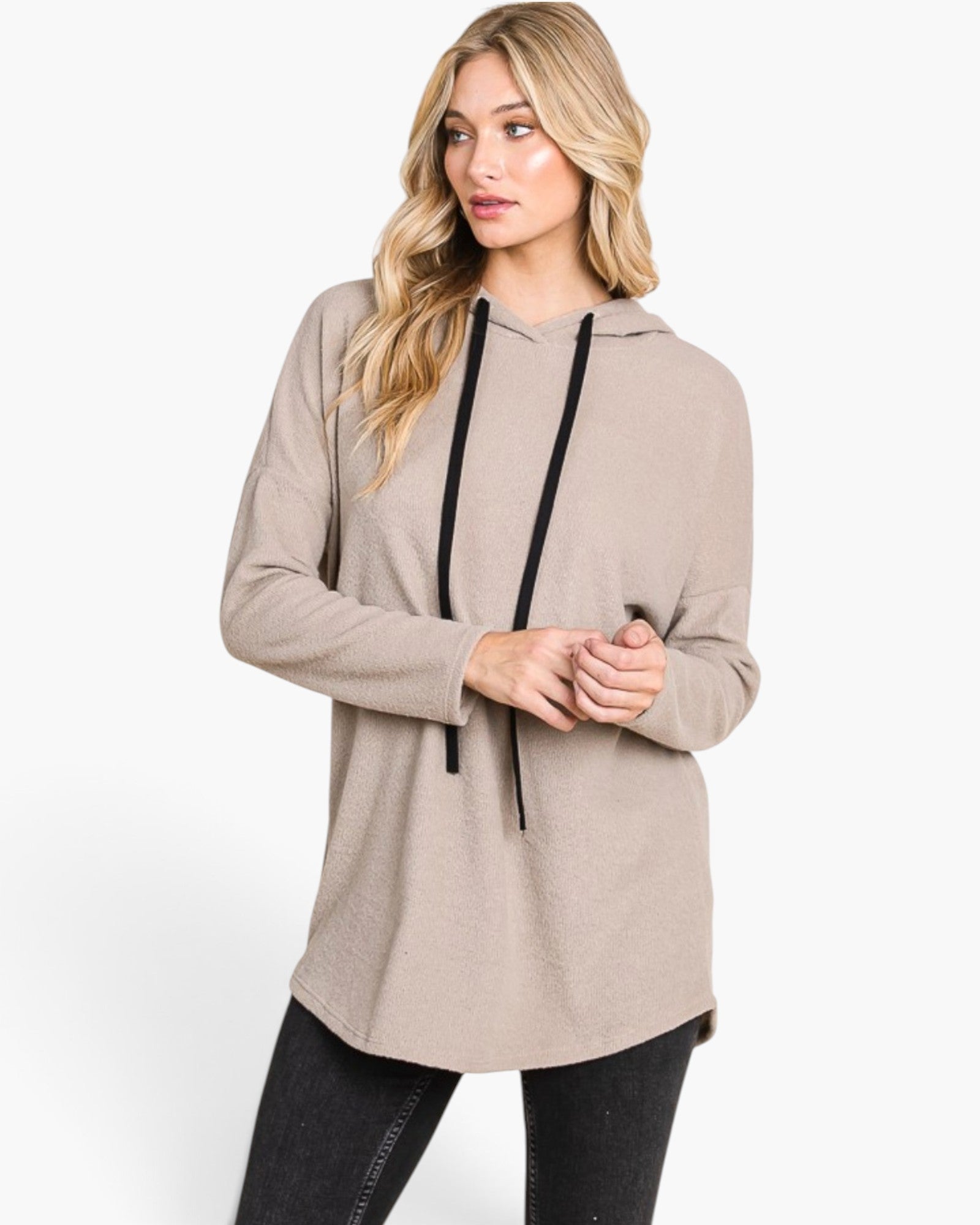 A woman modeling a light taupe-beige long-sleeve hooded tunic top with black drawstrings, paired with black leggings, against a plain white studio background.