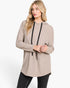 A woman modeling a light taupe-beige long-sleeve hooded tunic top with black drawstrings, paired with black leggings, against a plain white studio background.