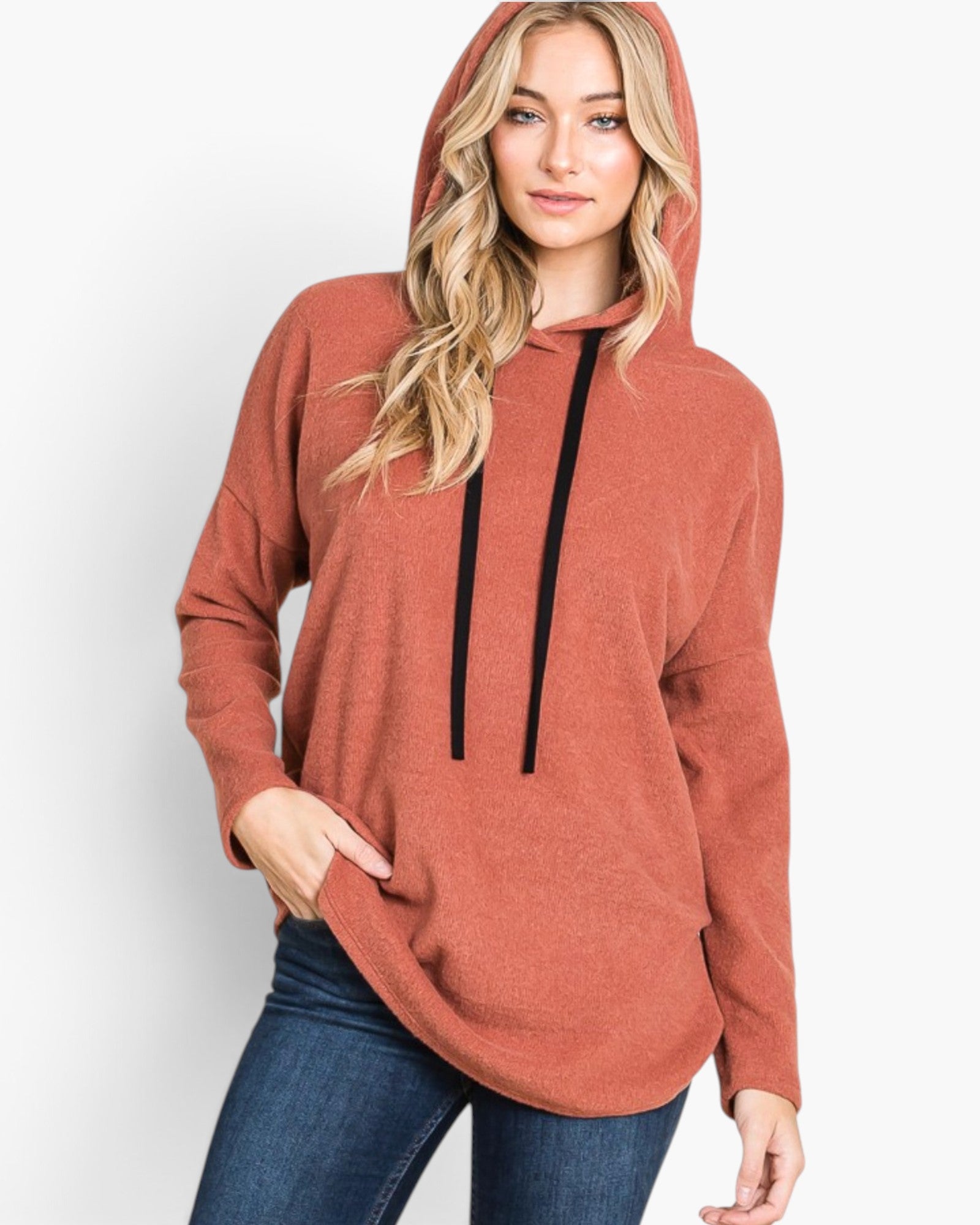 A woman wearing a rust-colored pullover hoodie with black drawstrings and blue jeans, standing against a plain white studio background.