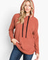 A woman wearing a rust-colored pullover hoodie with black drawstrings and blue jeans, standing against a plain white studio background.