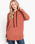 A woman wearing a heathered terracotta-colored pullover hoodie with black drawstrings, paired with blue jeans, against a plain white studio background.