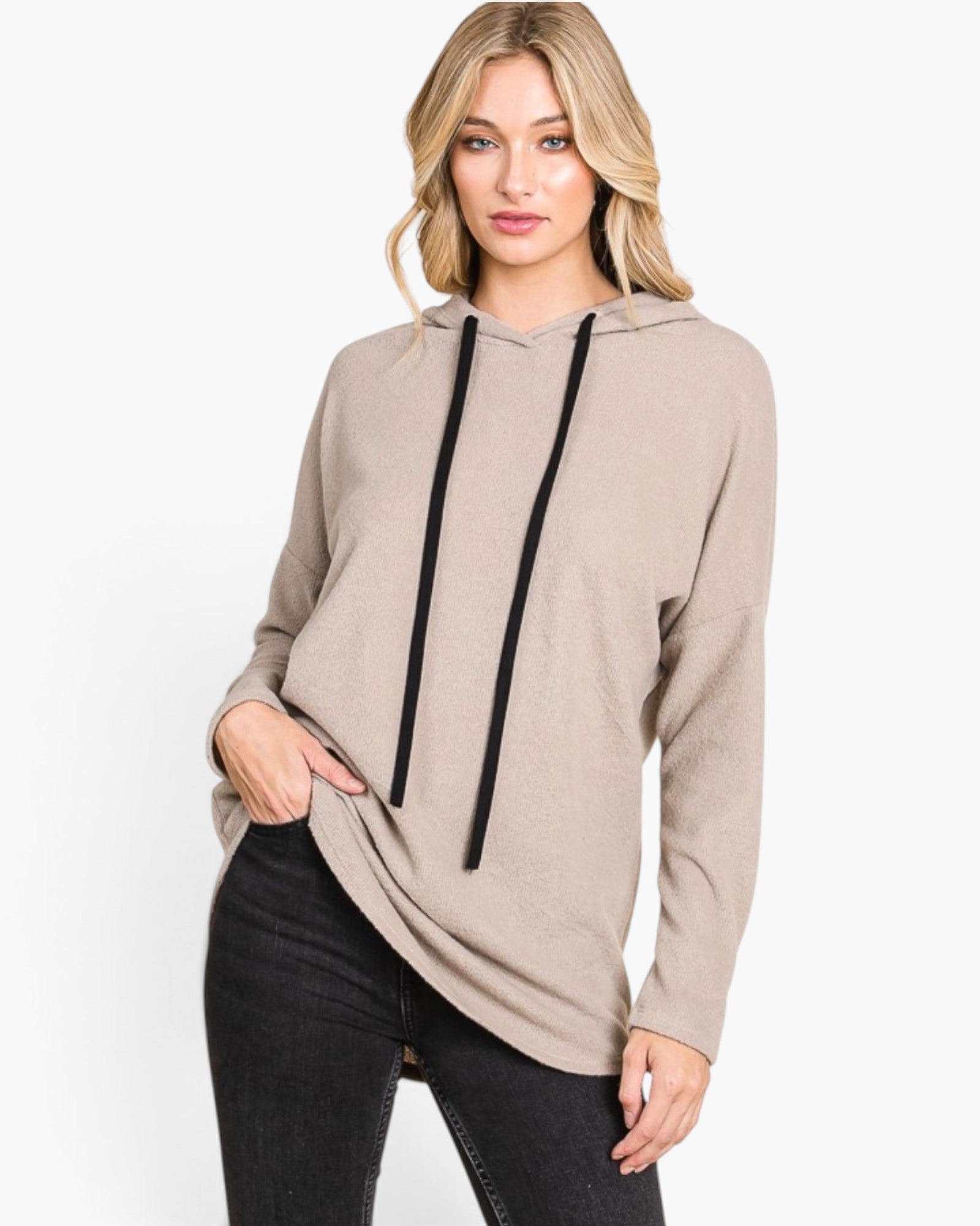 A woman modeling a light taupe-beige long-sleeve hoodie with black drawstrings, paired with black jeans, against a plain white studio background.
