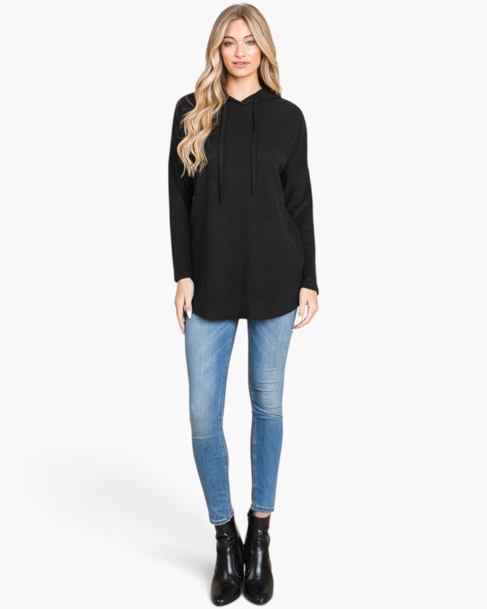 A woman modeling a black long-sleeve knit hoodie tunic with drawstrings, paired with blue skinny jeans and black ankle boots, against a plain white studio background.
