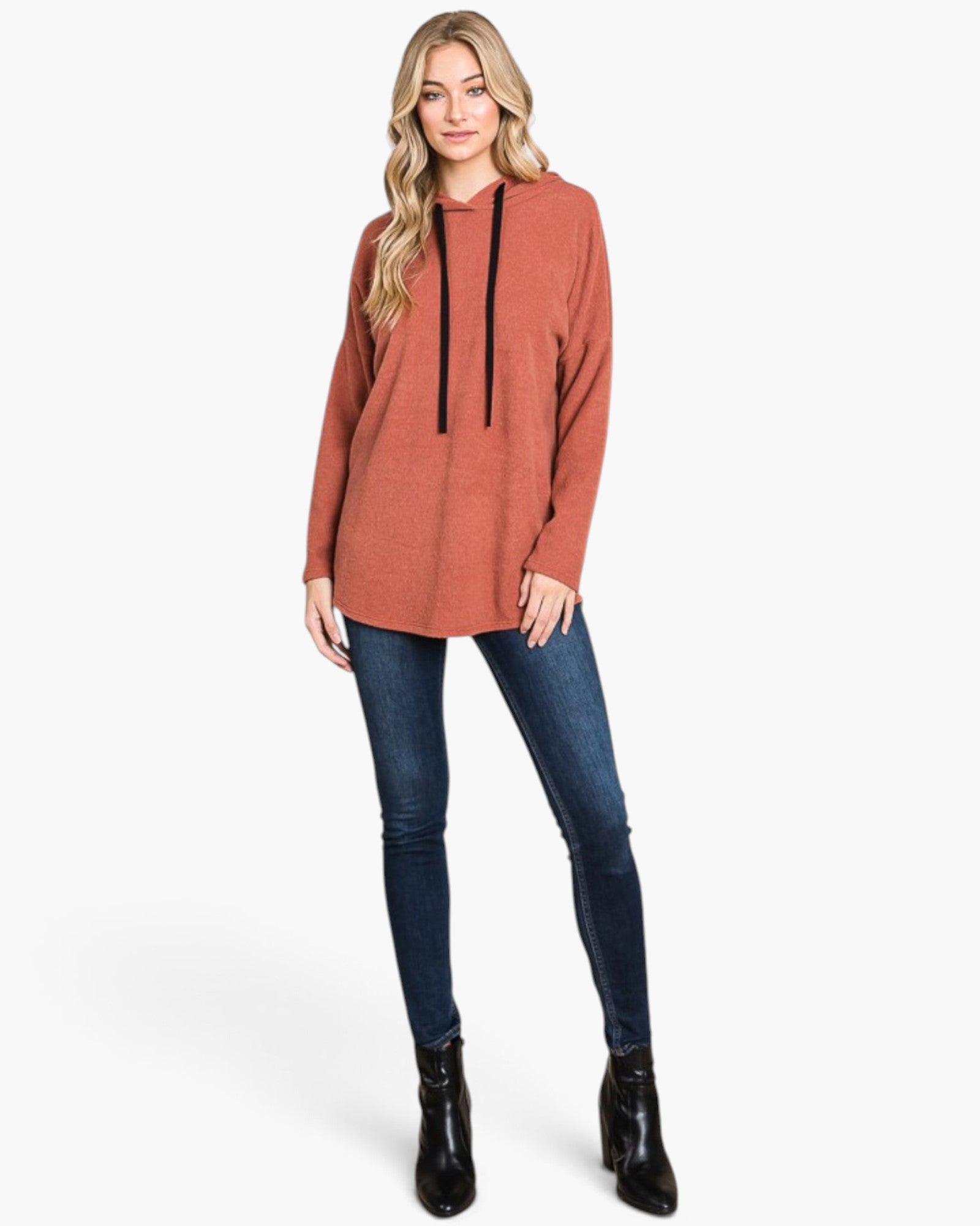 A woman modeling a terracotta-colored knit hoodie tunic with black drawstrings, paired with dark blue skinny jeans and black ankle boots, against a solid off-white background.