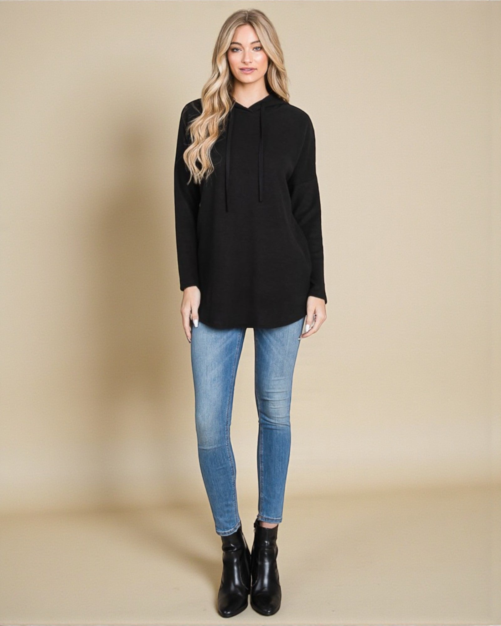 Woman wearing a black hoodie, blue jeans, and black boots on a beige background