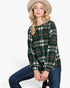 A woman wearing a green plaid long-sleeve top with red and white stripes, blue jeans, and a beige hat, sitting against a plain white background.