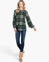 A woman modeling a green plaid long-sleeve top with blue skinny jeans and tan ankle boots, standing against a light gray studio background.