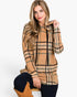 A woman wearing a tan and black plaid hooded sweatshirt, paired with dark pants and tan boots, posing against a light gray studio background.