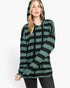 A woman wearing a green and black horizontally striped long-sleeve hoodie with a drawstring hood, paired with dark gray jeans, against a plain white studio background.
