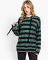 A woman modeling a green and black horizontally striped long-sleeve hoodie with black drawstrings, paired with dark gray jeans, against a plain white studio background.