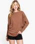 A woman modeling a brown knit sweater with a boat neckline and long sleeves, paired with black shorts, against a plain white background.