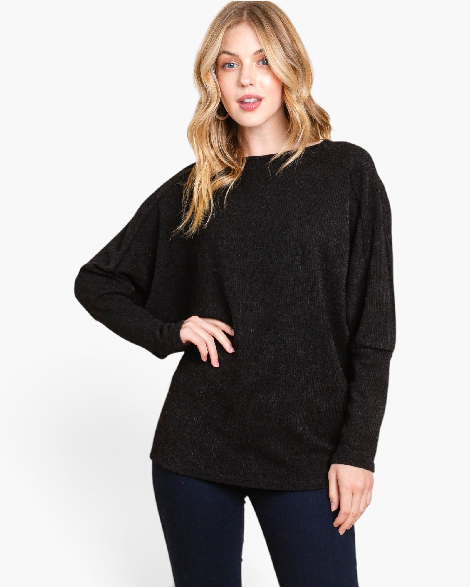 A woman modeling a black long-sleeve knit sweater with a relaxed, oversized fit and batwing sleeves, paired with dark blue jeans, against a plain white studio background.