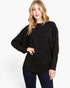 A woman modeling a black long-sleeve knit sweater with a relaxed, oversized fit and batwing sleeves, paired with dark blue jeans, against a plain white studio background.
