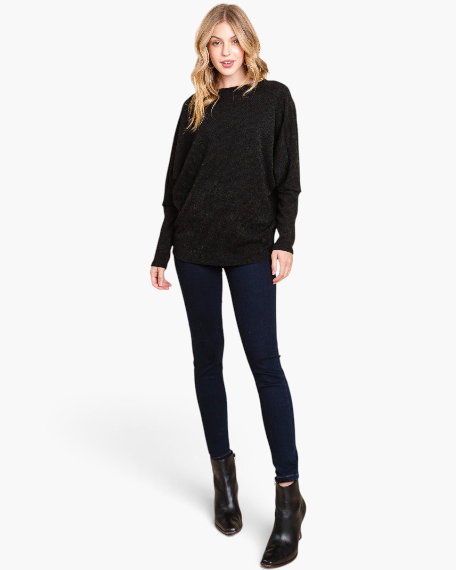 A woman modeling a black long-sleeve knit sweater, dark navy skinny jeans, and black ankle boots against a light gray studio background.