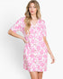 A woman modeling a short-sleeve button-front dress with a pink floral print on a white background, photographed against a clean light gray studio backdrop.
