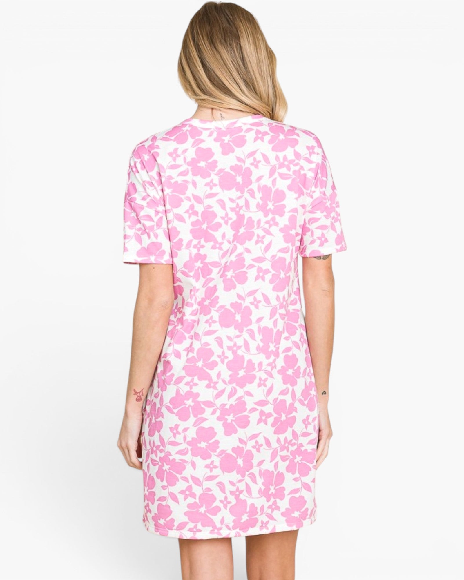 A woman viewed from the back wearing a short-sleeve shift dress with a pink floral pattern on a white background, set against a plain white studio backdrop.