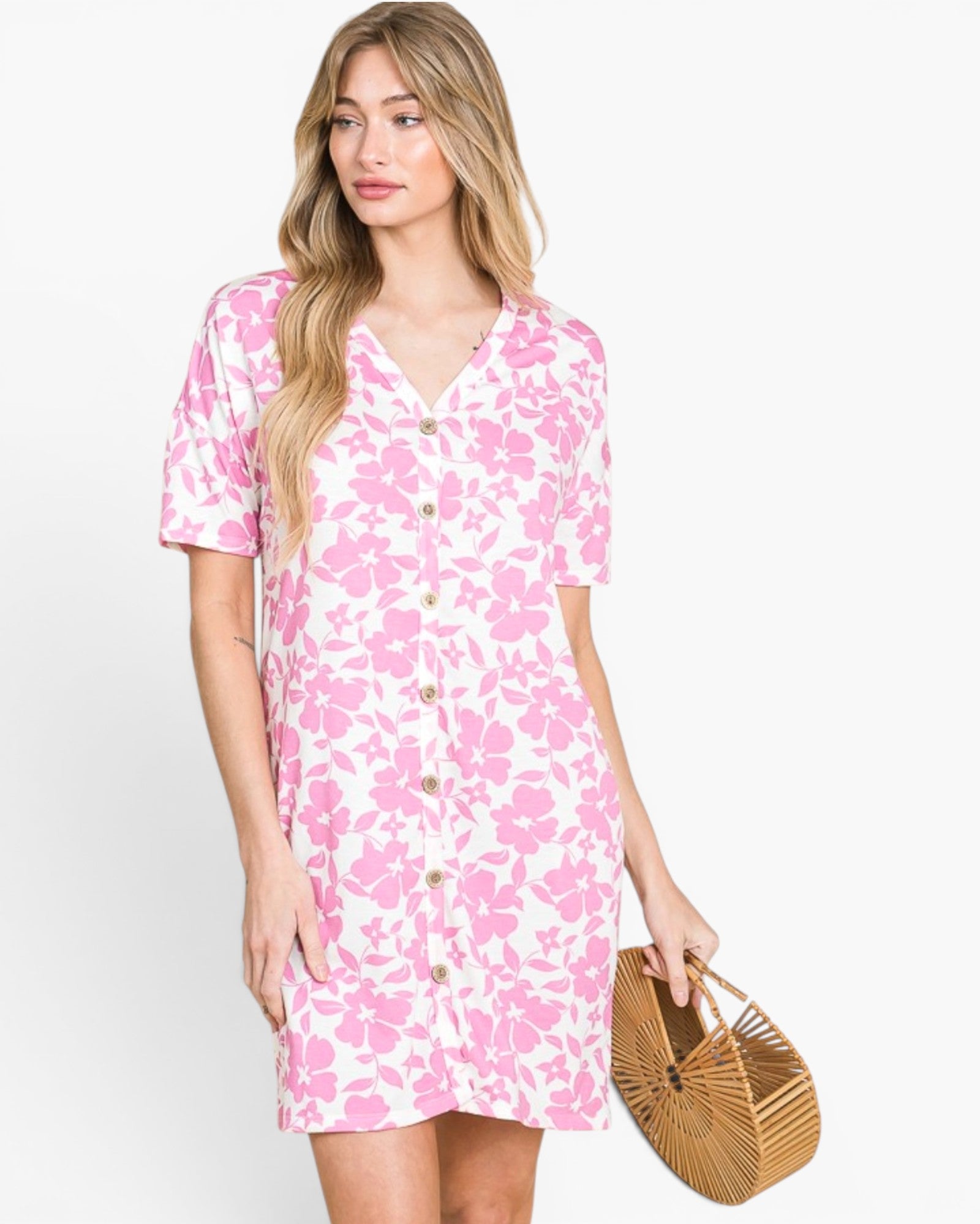 A woman modeling a short-sleeve V-neck button-front dress with a pink floral print on a white background, holding a round woven straw bag against a plain light gray studio background.