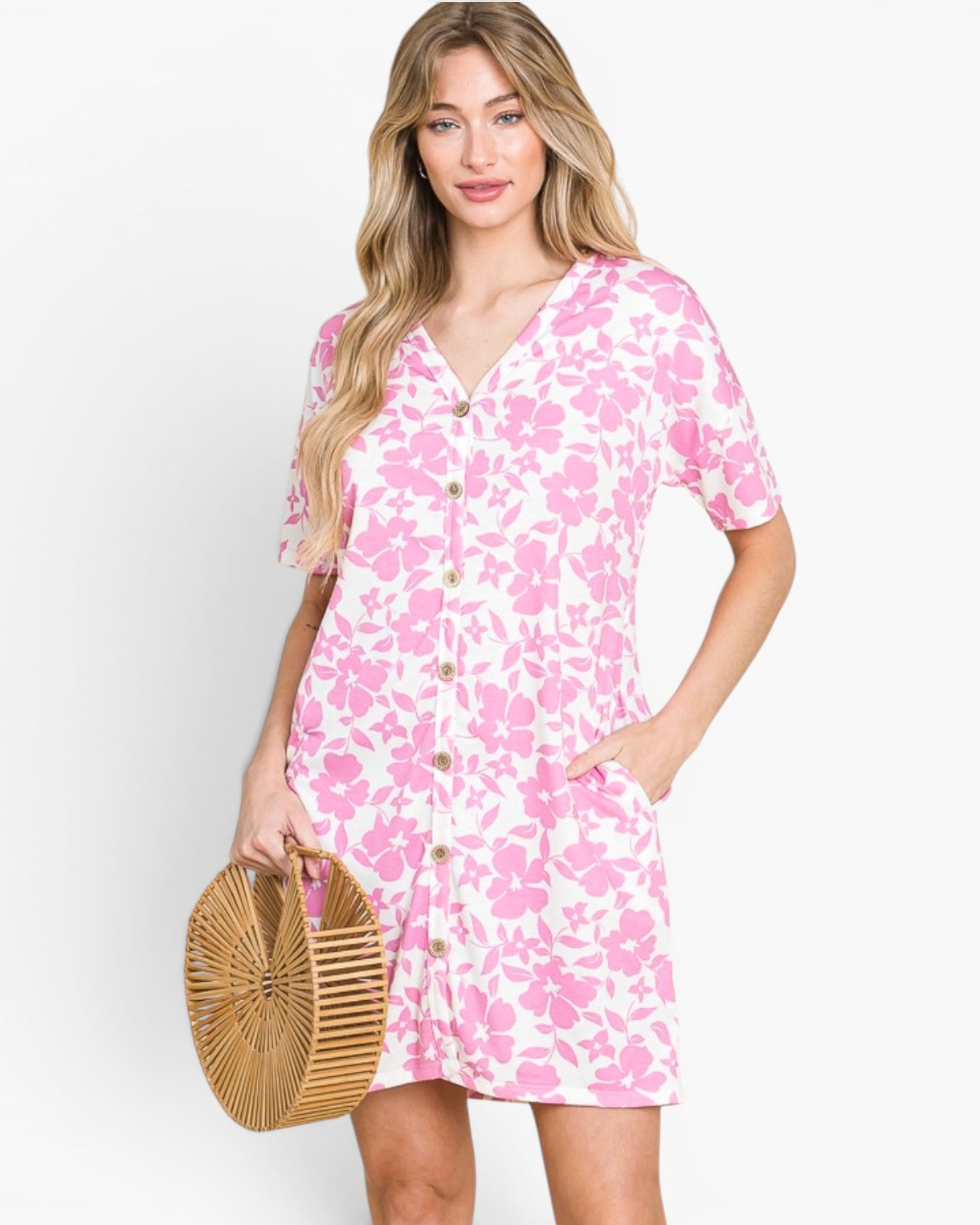 A woman modeling a short-sleeve pink and white floral print mini dress with a button-down front, holding a round woven straw bag against a light gray studio background.