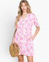A woman modeling a short-sleeve pink and white floral print mini dress with a button-down front, holding a round woven straw bag against a light gray studio background.