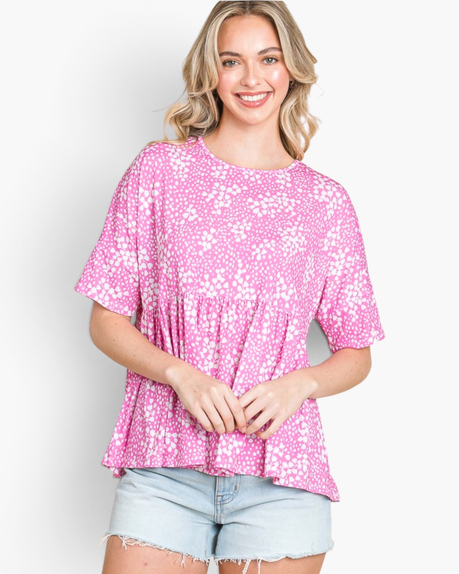 A woman wearing a pink and white floral print short-sleeve peplum top and light blue denim shorts, standing against a plain white studio background.