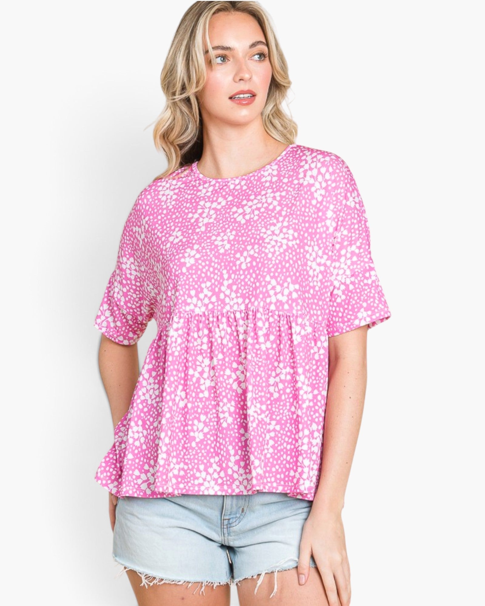 A woman wearing a pink short-sleeve peplum top with a white floral pattern, paired with light-wash denim shorts, against a plain white studio background.