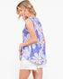 A woman wearing a sleeveless lavender top with a large white and purple daisy floral print, paired with white denim shorts, shown from a side-back angle against a plain white background.