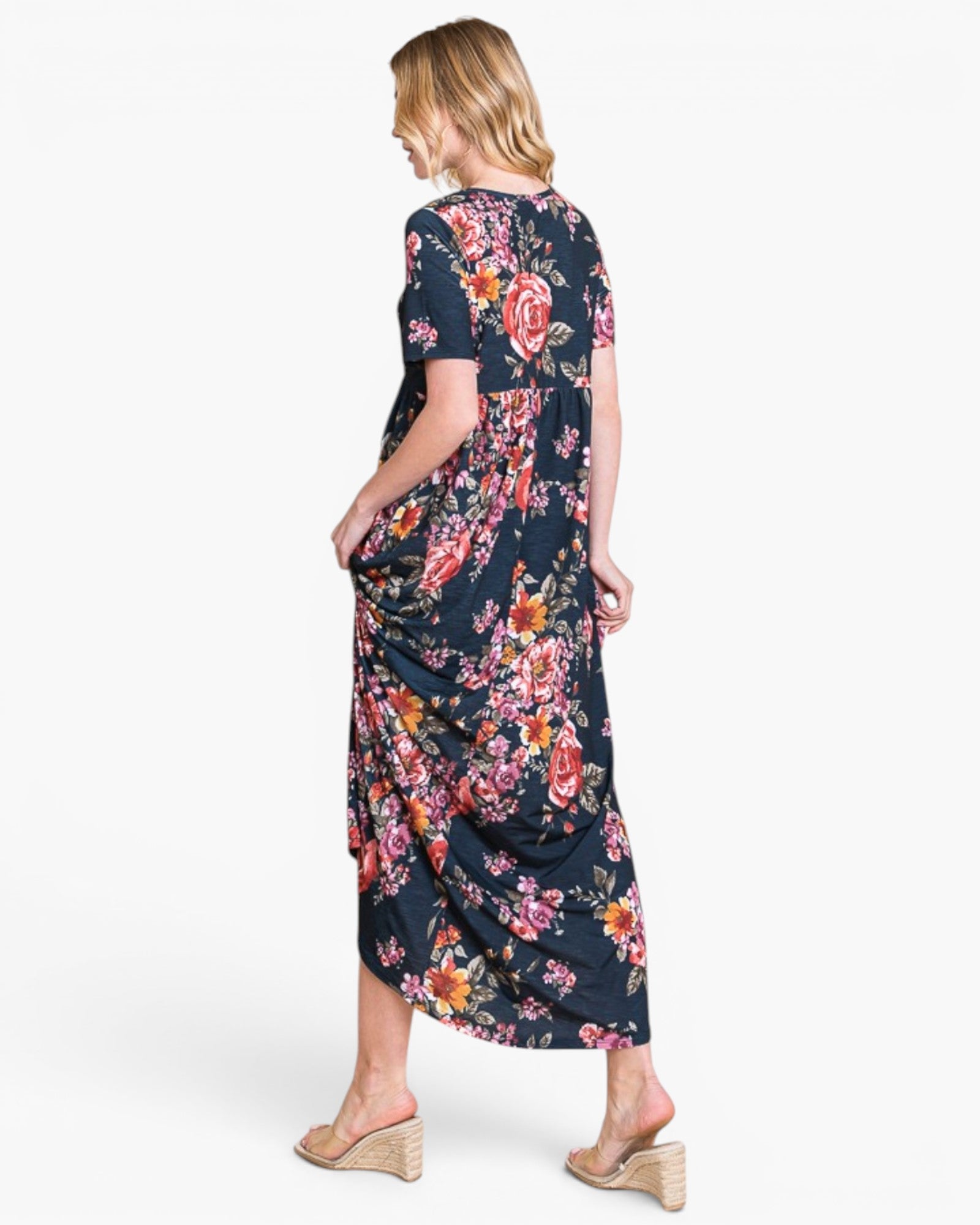 A woman modeling a short-sleeve midi dress with a dark navy blue background and a vibrant multicolor floral print, shown from a rear three-quarter view against a plain white studio background.