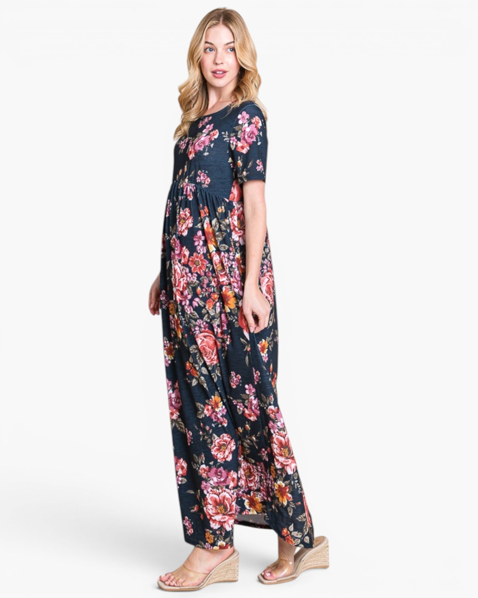 A woman modeling a short-sleeve maxi dress with a dark navy blue background and a vibrant pink, red, and orange floral print, paired with beige wedge sandals against a plain white studio background.