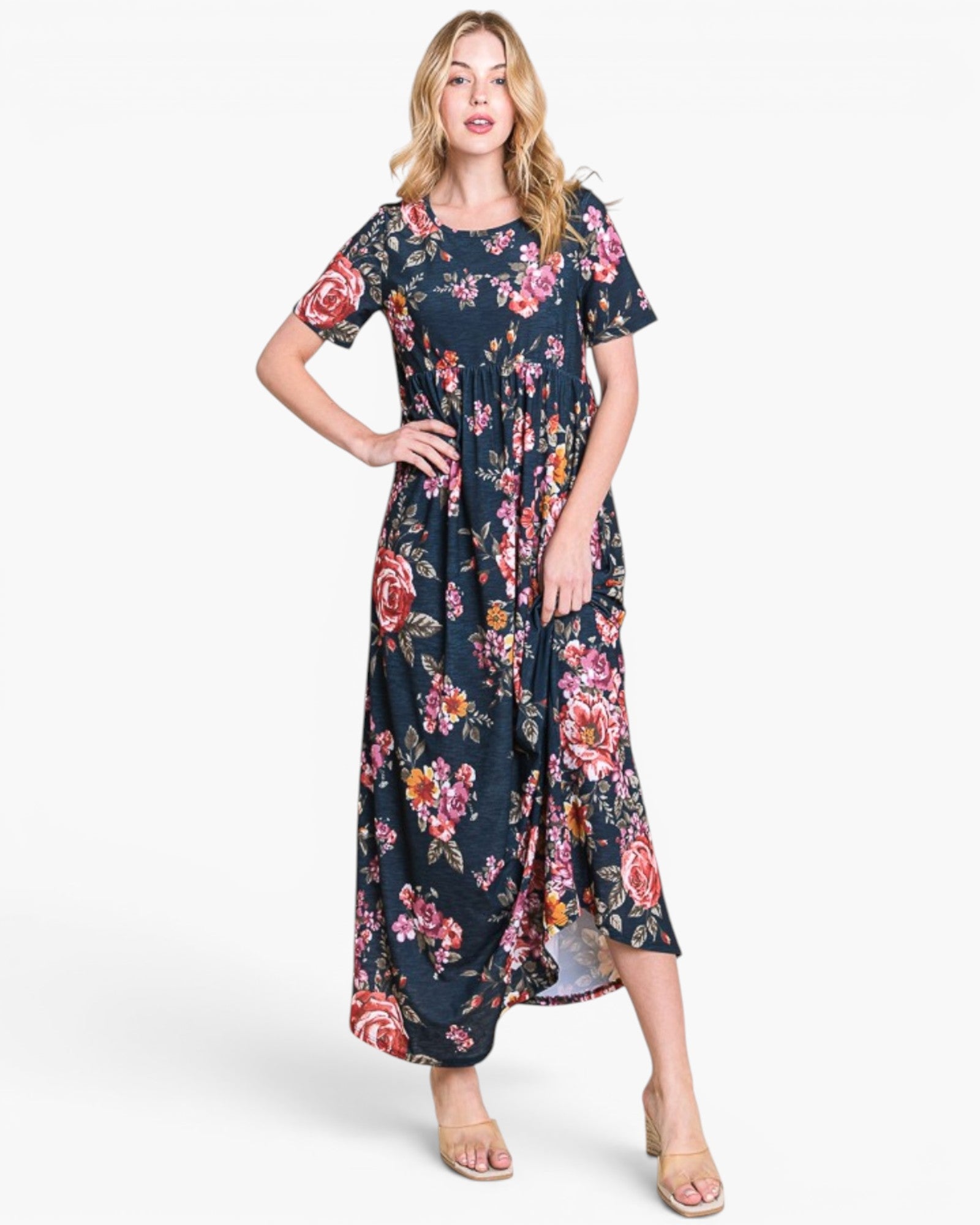A woman modeling a short-sleeve, midi-length floral dress with a dark navy base and pink, red, and yellow floral print, paired with beige heeled sandals against a light gray studio background.