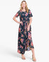 A woman modeling a short-sleeve, midi-length floral dress with a dark navy base and pink, red, and yellow floral print, paired with beige heeled sandals against a light gray studio background.