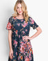 A woman modeling a short-sleeve navy blue dress with a vibrant pink, red, and orange floral print against a light gray studio background.