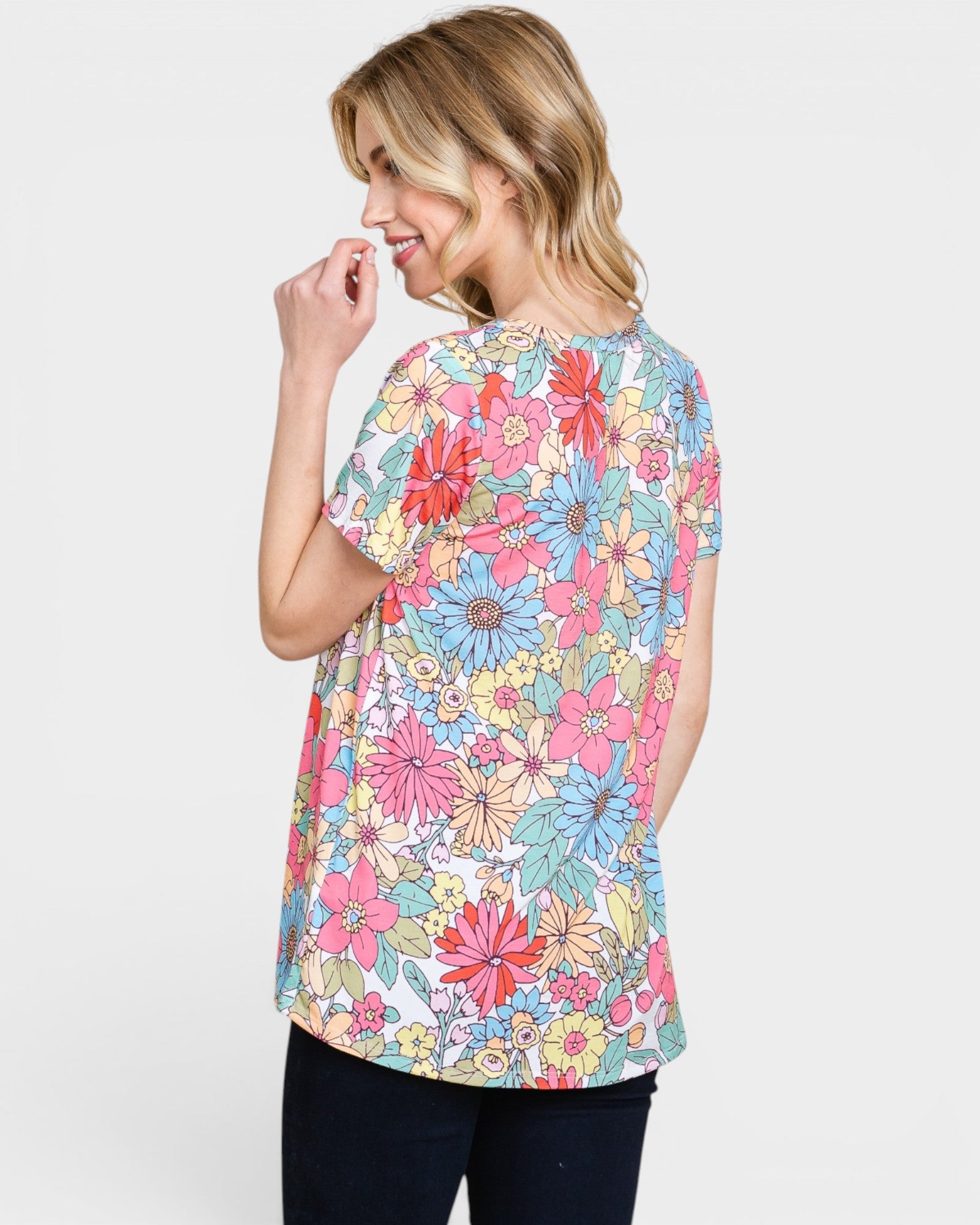 A woman wearing a short-sleeve tunic top with a colorful floral print, shown from a three-quarters back view against a light gray background.