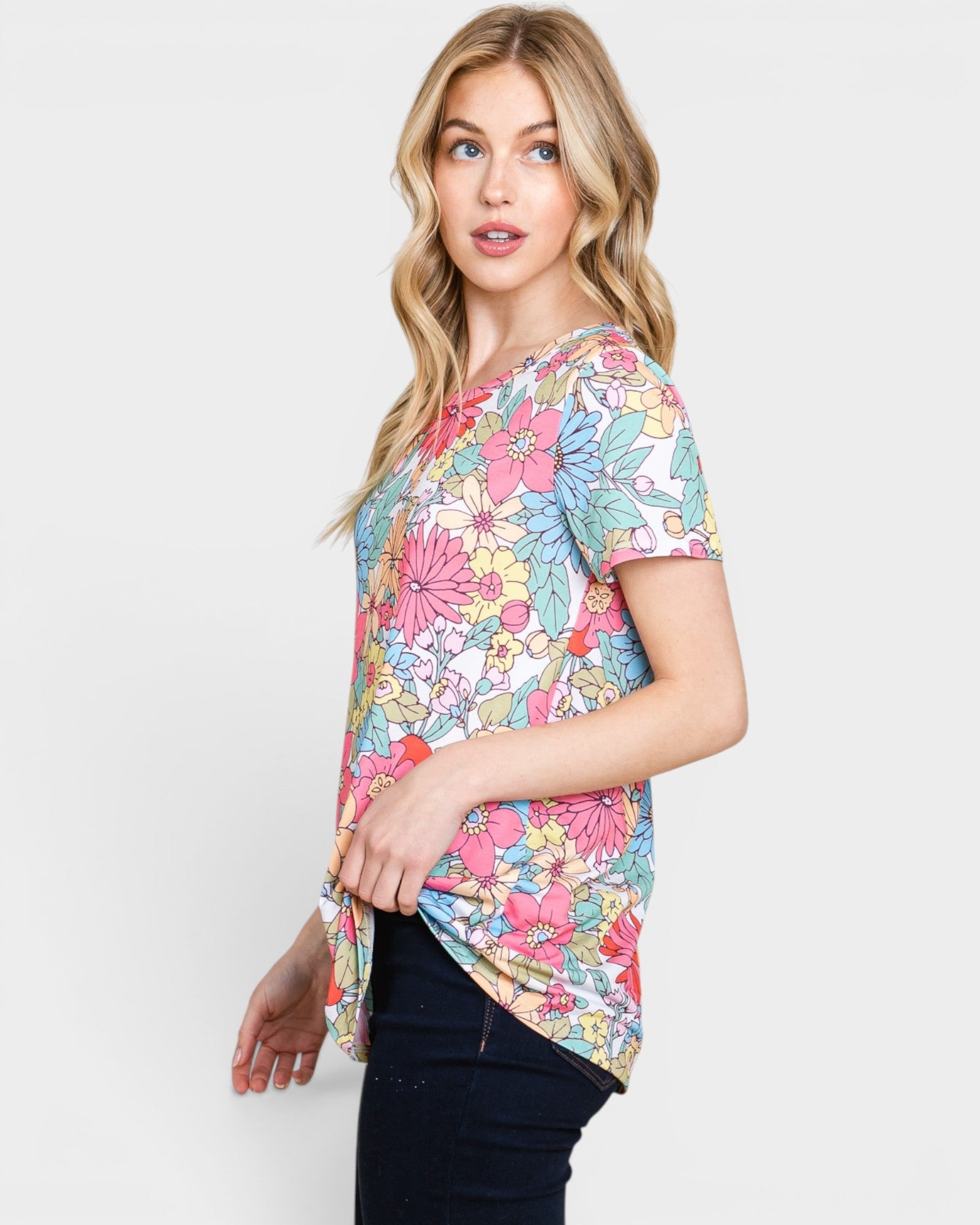 A woman modeling a short-sleeve V-neck top with a colorful floral and leaf print in pink, blue, yellow, and green on a white base, paired with dark jeans against a light gray studio background.