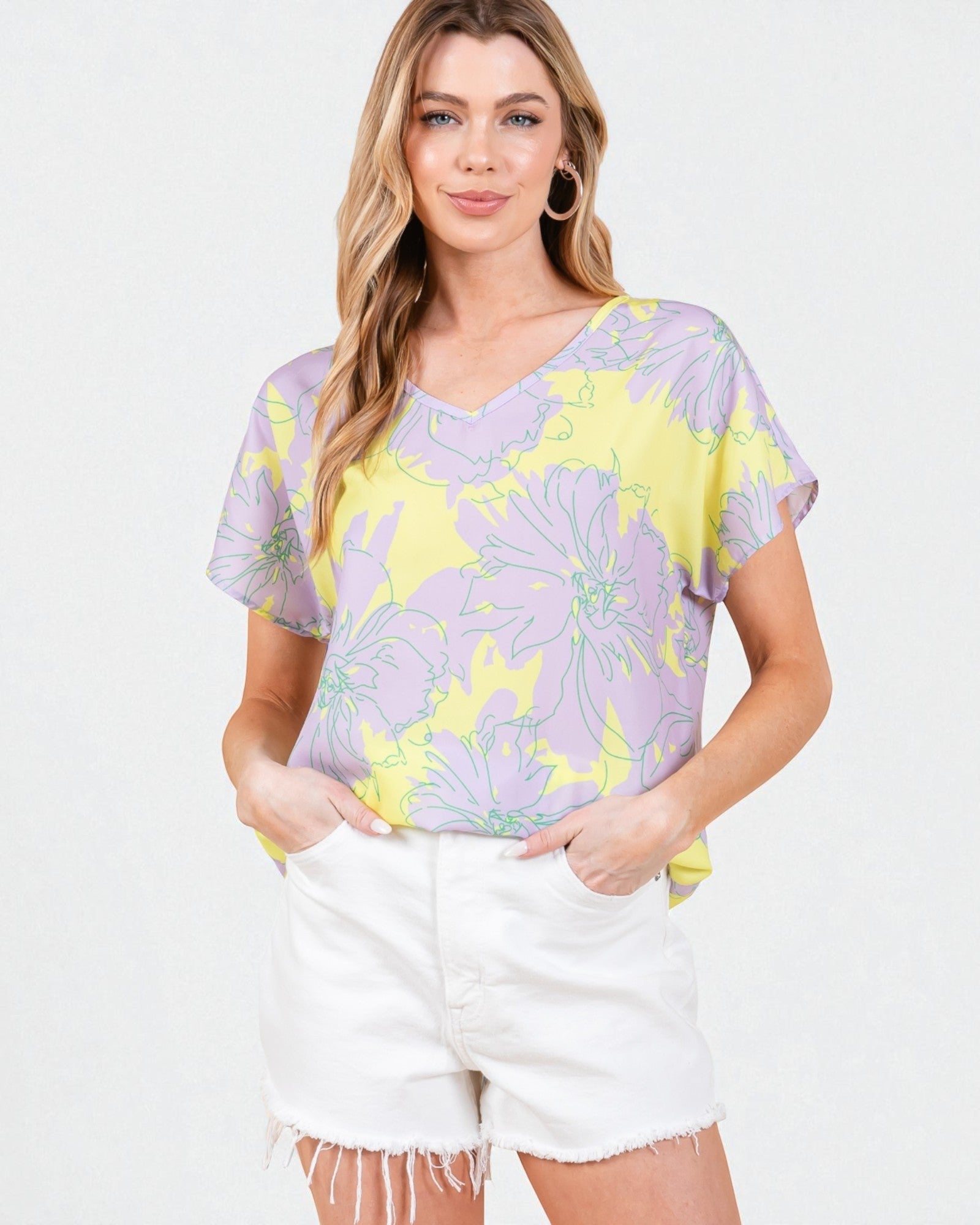 Woman wearing a purple and yellow floral top with white shorts on a light gray background