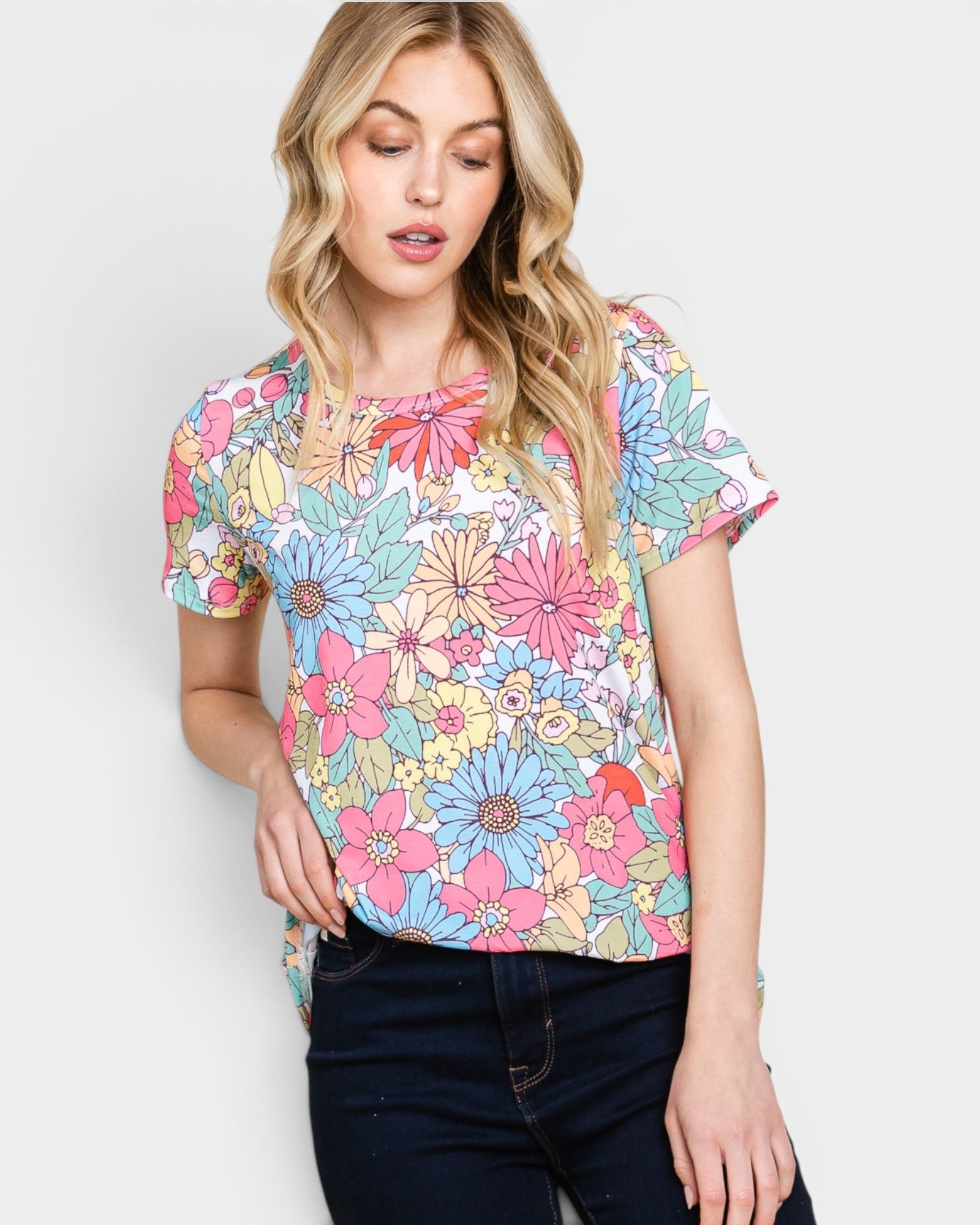 A woman wearing a short-sleeve crewneck top with a colorful floral print in pink, blue, yellow, and green on a white background, paired with dark blue jeans, against a light gray studio background.