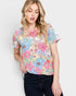 A woman wearing a short-sleeve crewneck top with a colorful floral print in pink, blue, yellow, and green on a white background, paired with dark blue jeans, against a light gray studio background.
