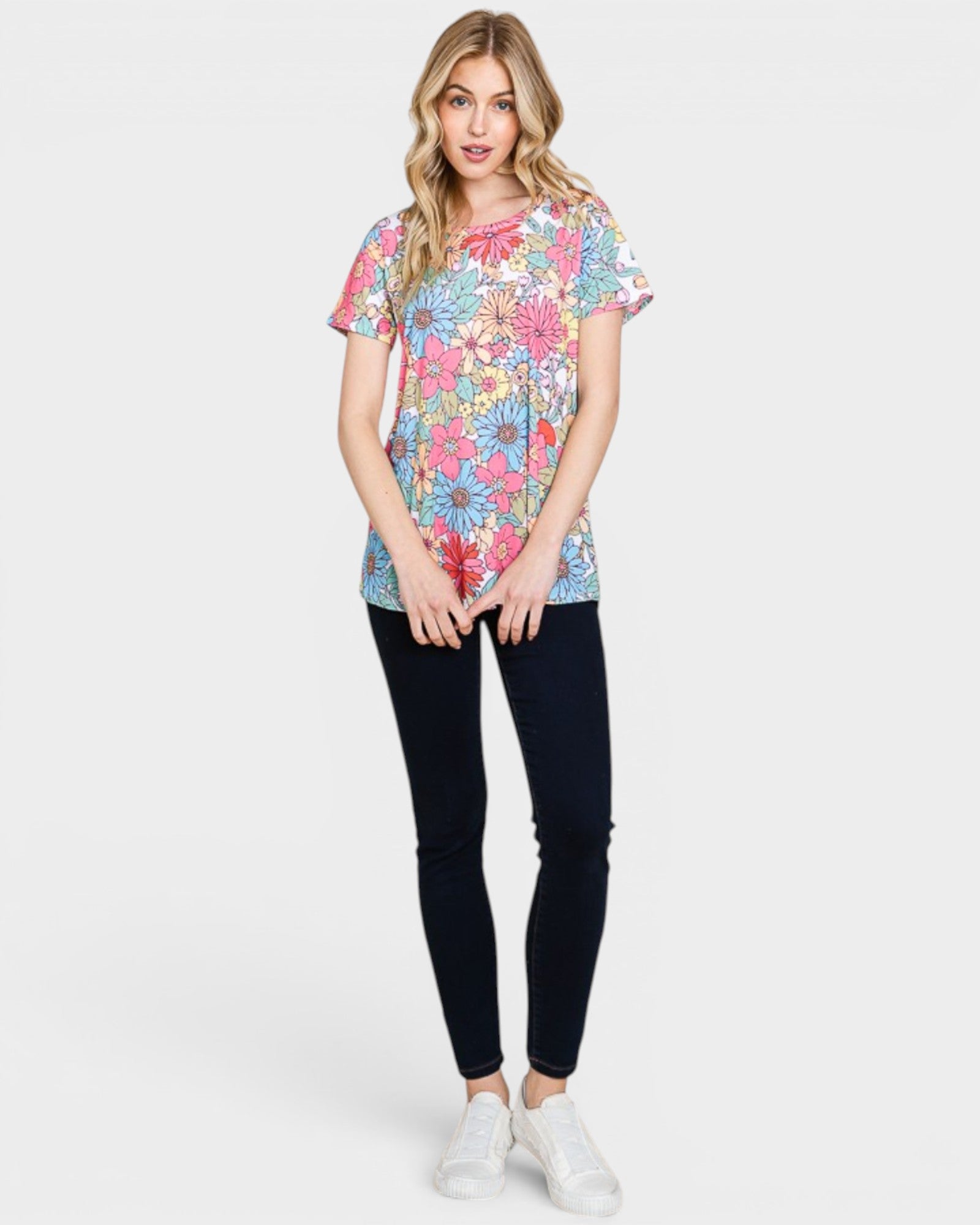 A woman modeling a short-sleeve crewneck t-shirt with a vibrant multicolor floral pattern, paired with black skinny jeans and white sneakers, against a light gray studio background.
