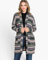 A woman modeling a long open-front cardigan with a multicolor geometric Fair Isle pattern in black, green, pink, and white, worn over a black top and blue jeans against a plain white studio background.