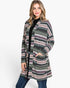 A woman modeling a long open-front cardigan with a multicolor Fair Isle pattern in green, pink, beige, and black, worn over a black top and blue jeans against a plain white background.