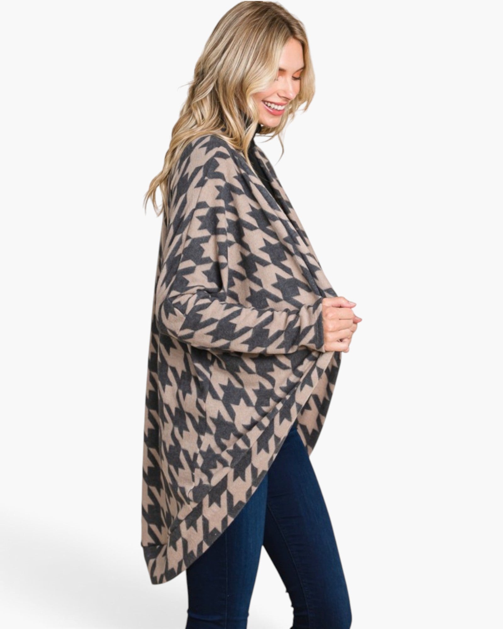 A woman wearing a beige and black houndstooth patterned open-front cardigan over a black top and dark blue jeans, photographed against a clean white studio background.