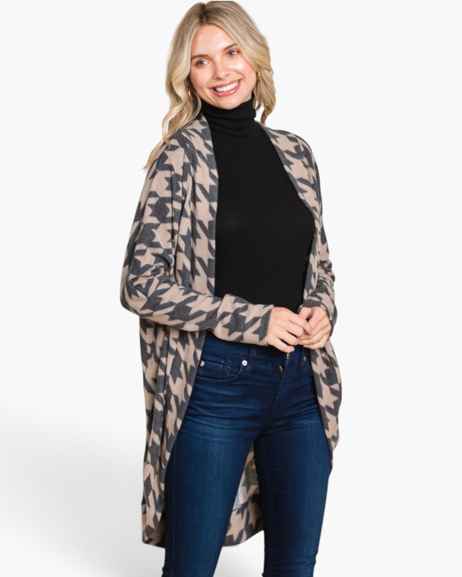 A woman modeling a beige and black houndstooth patterned open-front cardigan over a black turtleneck and dark blue jeans, photographed against a plain white studio background.