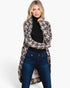 A woman modeling a beige and gray houndstooth patterned open-front cardigan over a black turtleneck and dark blue jeans, photographed against a clean white studio background.